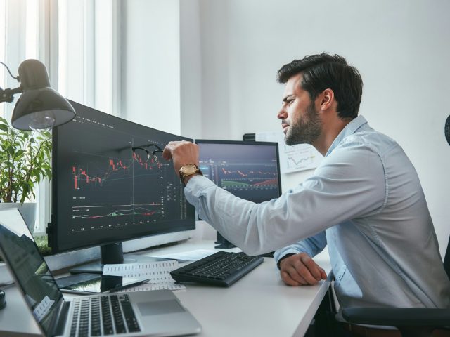 Analyzing data. Thoughtful young trader in eyeglasses is pointing at graphs on computer screen while working in his modern office. Stock exchange. Trade concept. Investment concept